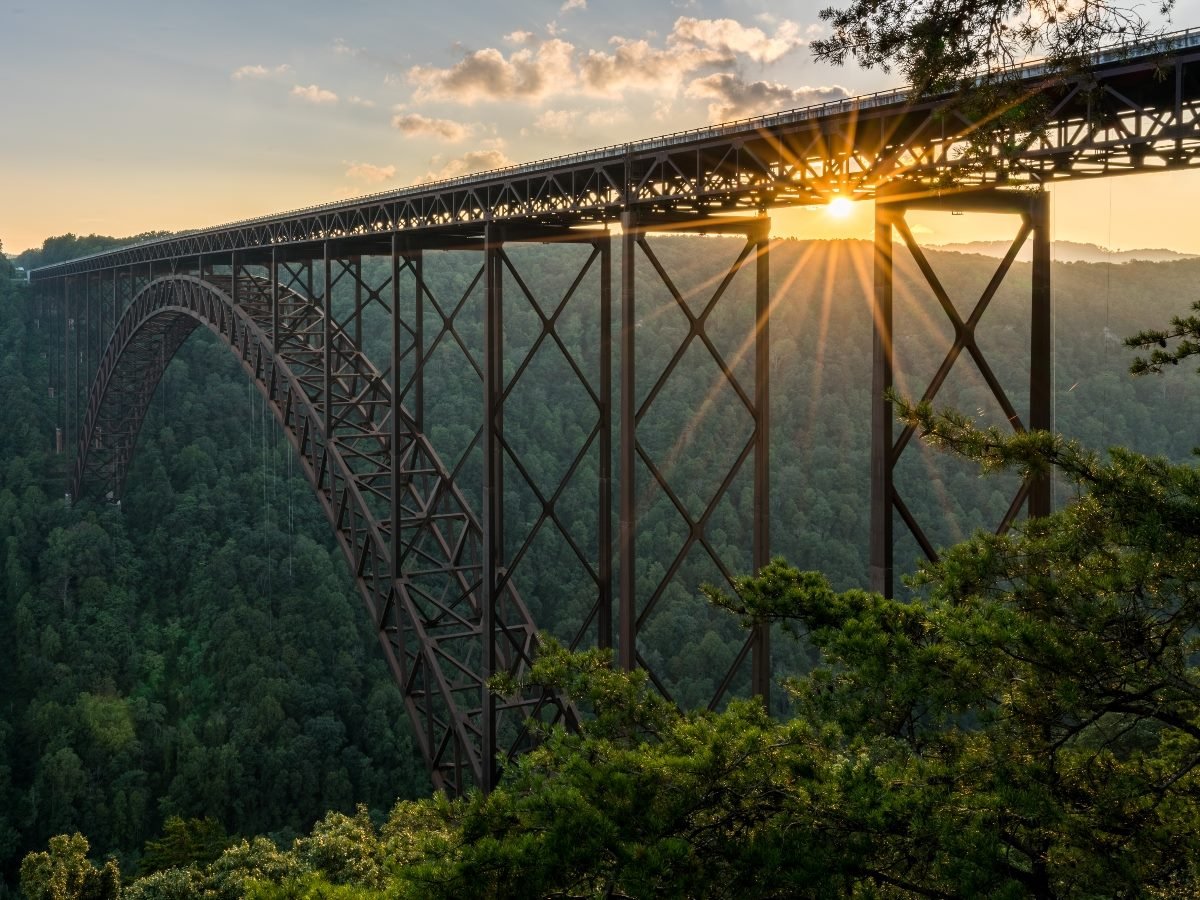 The New River Gorge Bridge in West Virginia at sunset, with sunlight streaming through the steel arch above the forested canyon.