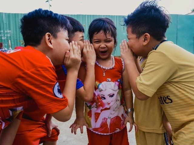 Four children gather around another child and tell secrets to her ears.
