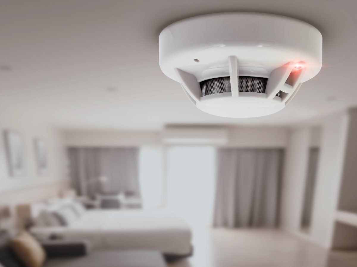 Close-up of a smoke detector with a red warning light activated inside a modern hotel room.
