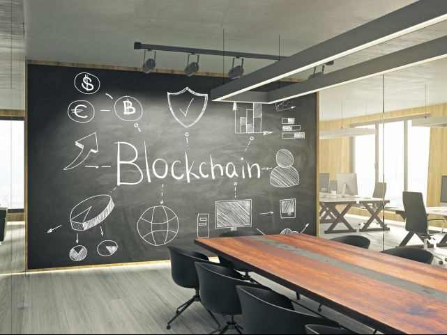 An open office setting with a large chalkboard in the middle. The chalkboard says blockchain - Blockchain in the workplace concept