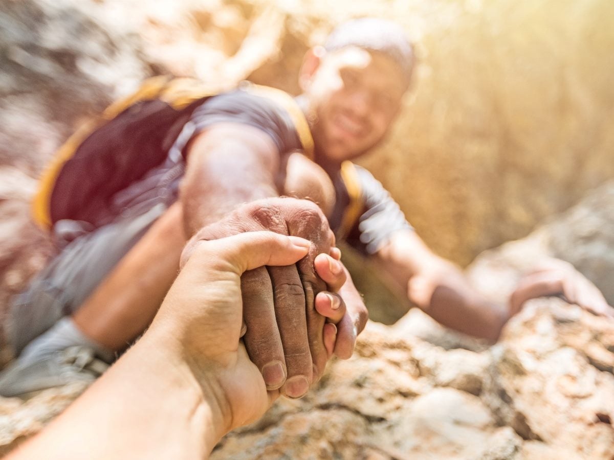 Close-up photo of one person pulling another climber upward by the hand on a rocky trail, symbolizing trust, support and reliability.