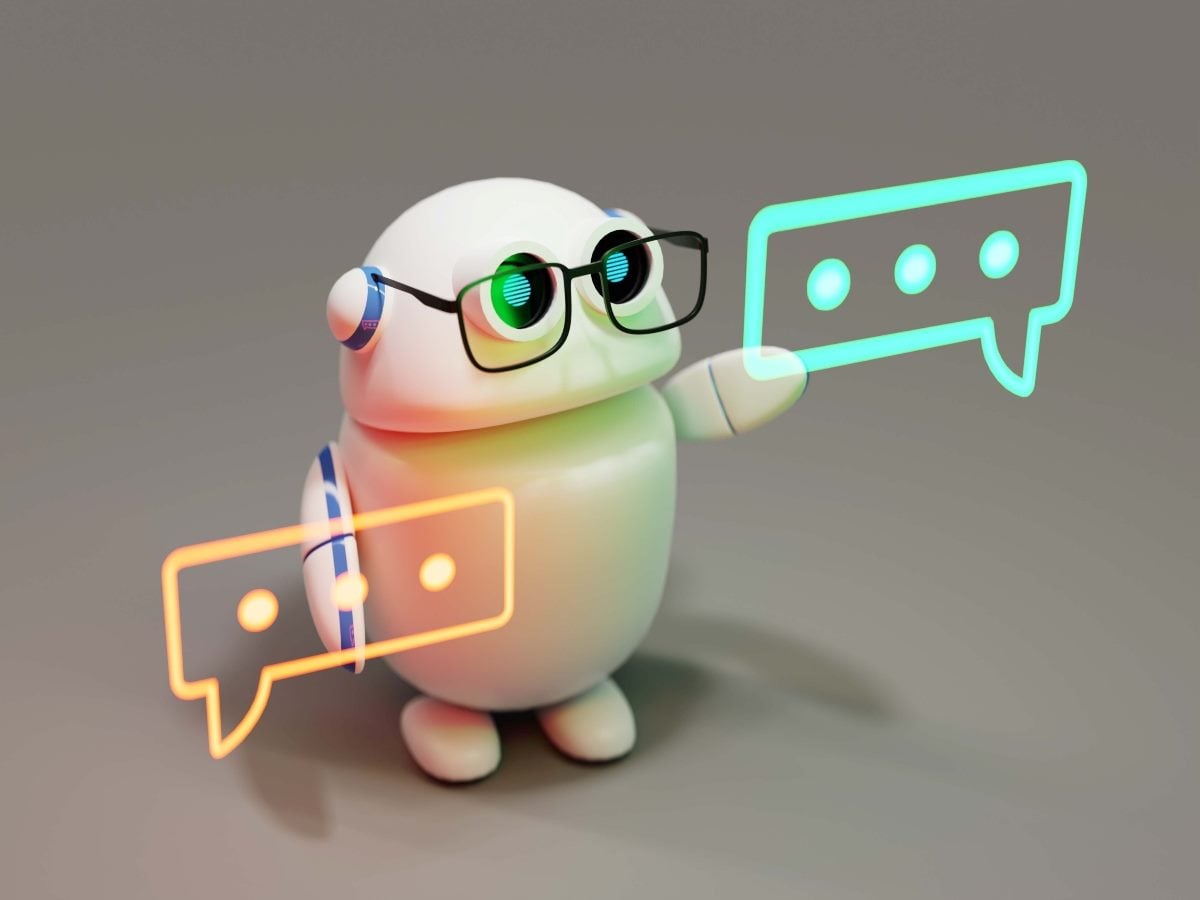 A small round white robot with a glossy, egg-shaped head and body stands facing slightly to the right against a plain gray background, wearing black rectangular glasses that frame its large round eyes, which glow green and blue with light reflections. The robot has short white arms and stubby feet, with a blue-and-white striped band circling one earlike side node on its head. It raises one arm toward a glowing teal speech-bubble outline containing three dots, positioned to the upper right, while a second glowing amber-orange speech-bubble outline, also with three dots, floats to the lower left near the robot's body. Soft ambient lighting casts a diffused shadow beneath the robot on the flat gray surface, giving the scene a clean, three-dimensional, softly lit studio look.