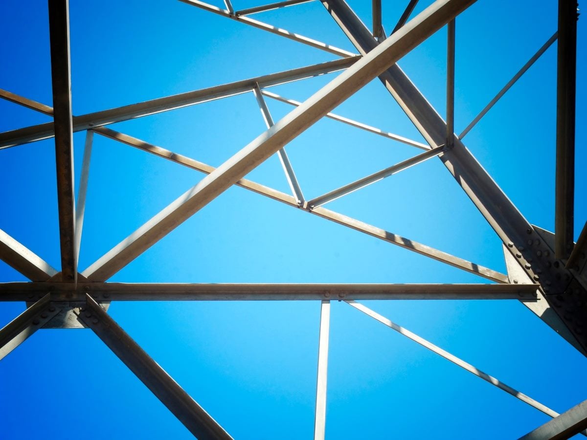 Abstract architectural photograph looking up through intersecting steel beams and support trusses against a bright blue sky, creating a geometric framework pattern that symbolizes structure, interconnected systems and foundational design.