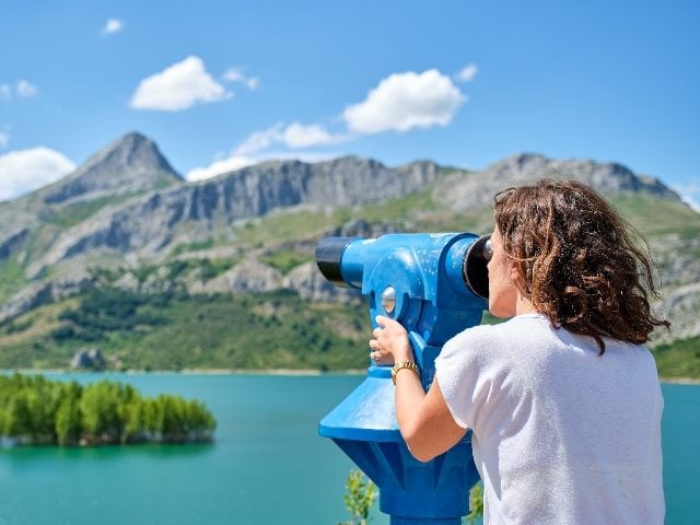 Woman with long curly hair looking through a telescope at a mountain landscape on a sunny day.