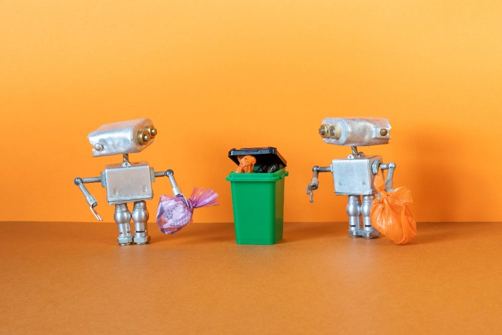 Two small silver metal robots are holding garbage bags ready to be disposed of in a green dumpster, trash can against an range background in piece about B2B marketing strategies.