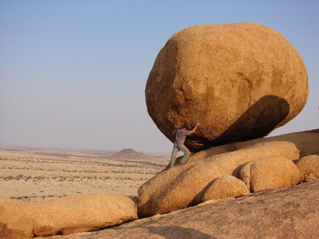 man standing beside rock formation and pretending to be holding it up