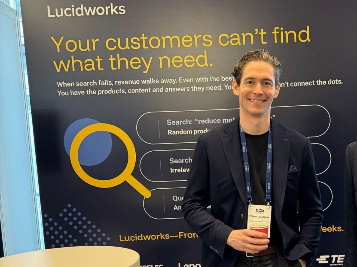 A man wearing a conference badge stands smiling in front of a Lucidworks booth backdrop that reads “Your customers can’t find what they need,” with graphics illustrating search challenges and a large yellow magnifying glass icon.