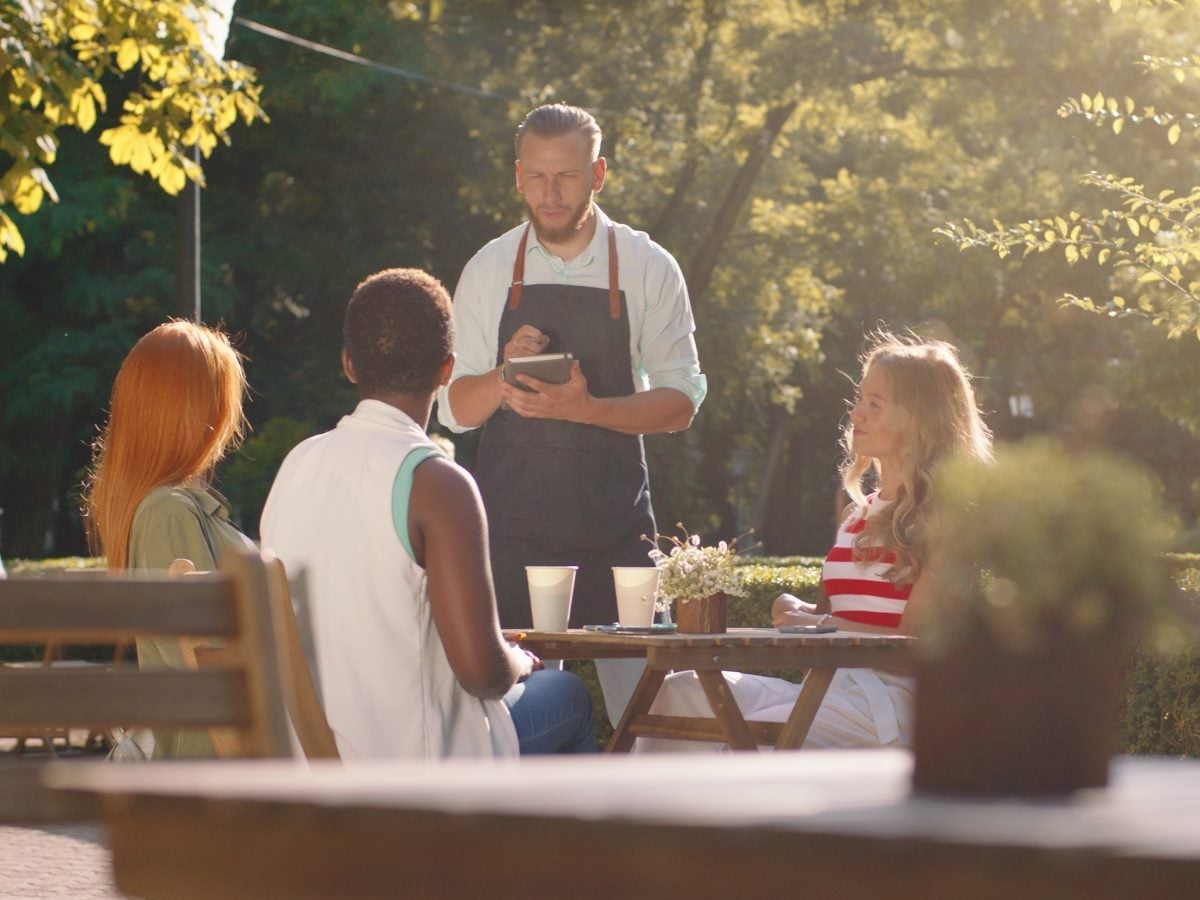In a forest scenery, a waiter with a beard and hair tied back is taking orders from a table with three women.