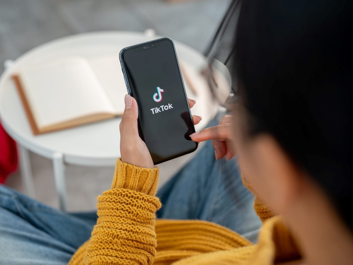 A person wearing a mustard-yellow sweater sits at a small white table holding a smartphone displaying the TikTok app logo on its screen. An open notebook rests on the table in the background, while the user's face is partially out of focus as they tap the phone.