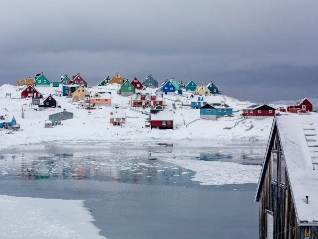 Snowy day in Greenland showing homes covered in snow across a body of water.
