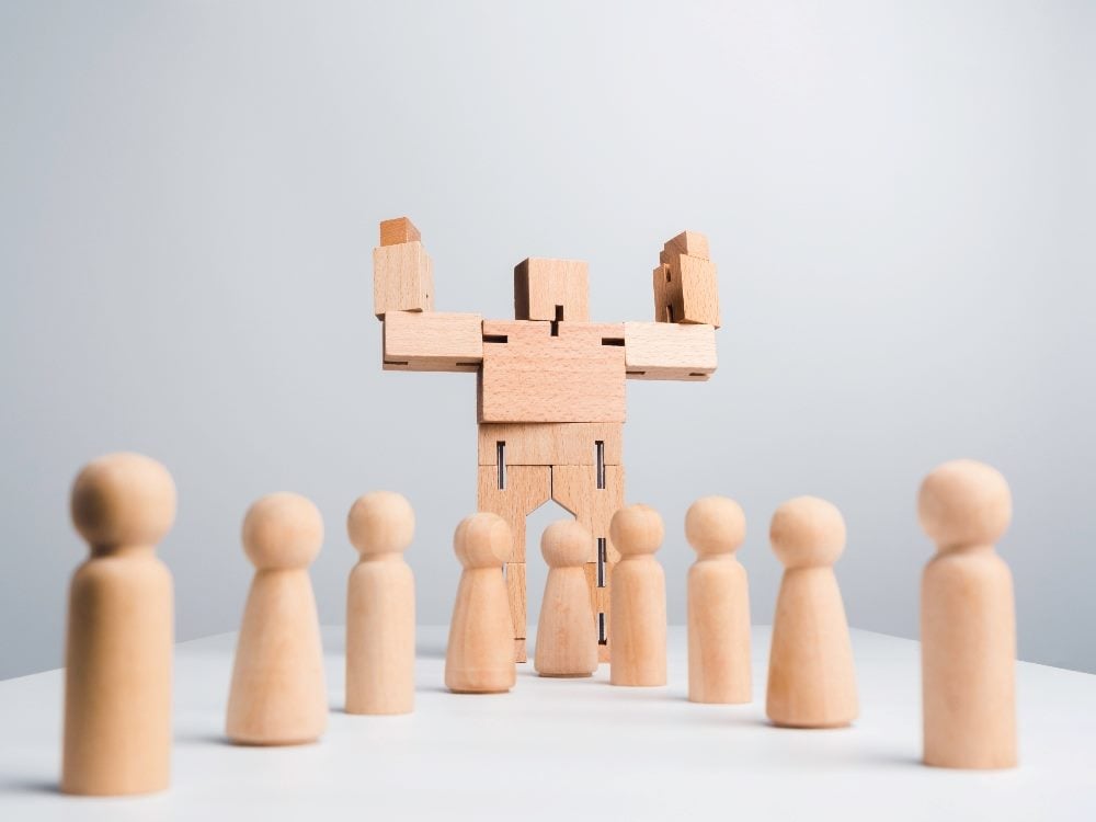 A wooden robot lifts its arms to show its muscles while standing among wooden human follower figures on white background in piece about AI in affiliate and influencer marketing.