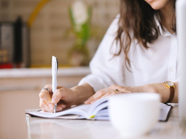 A. person with long dark hair sits at a kitchen table with a notebook handwriting a list with a blurred background of the room in piece about marketing leadership. 