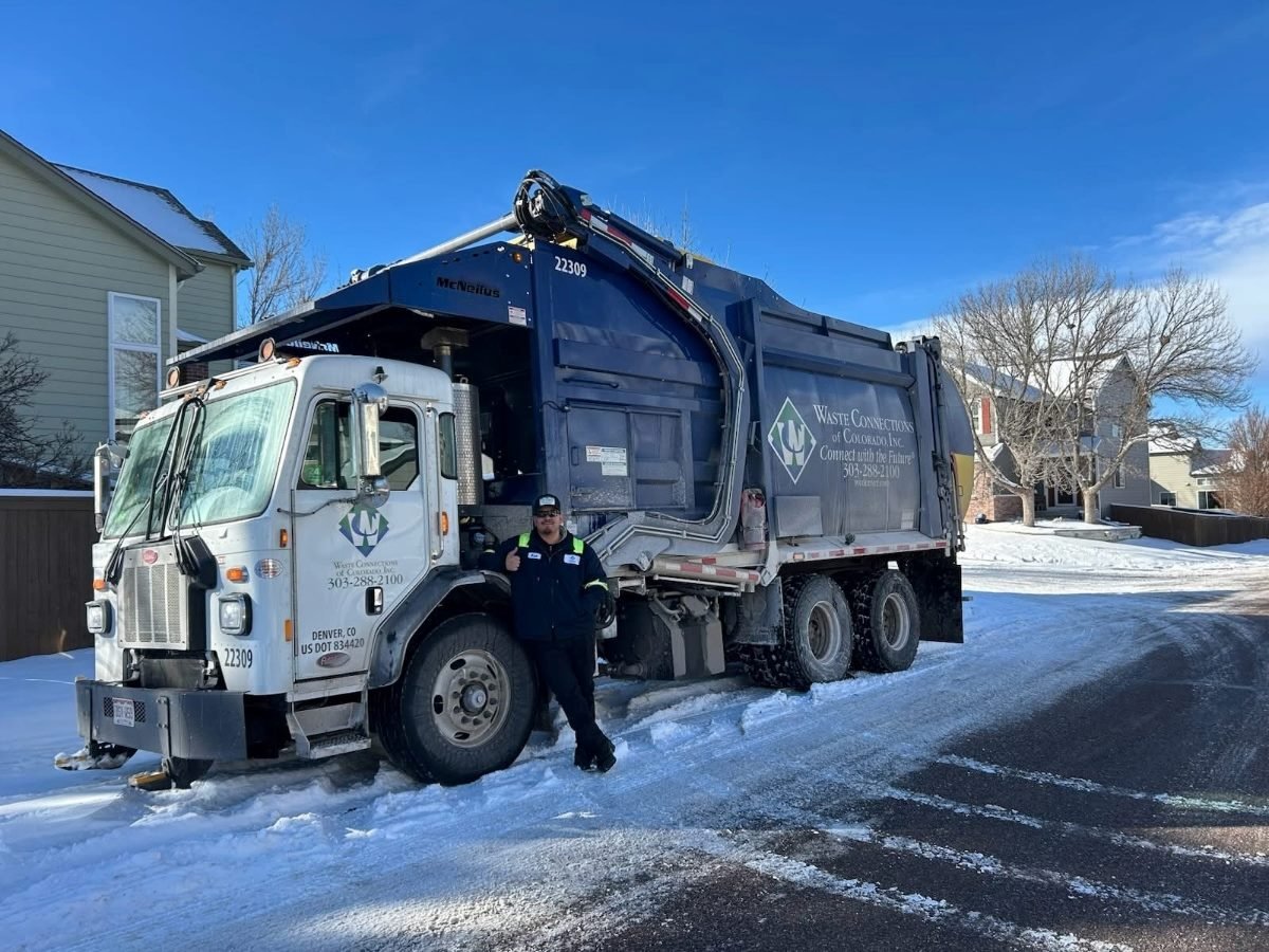 A Waste Connections garbage truck parked on a snowy residential street, with a sanitation worker standing beside it giving a thumbs-up.