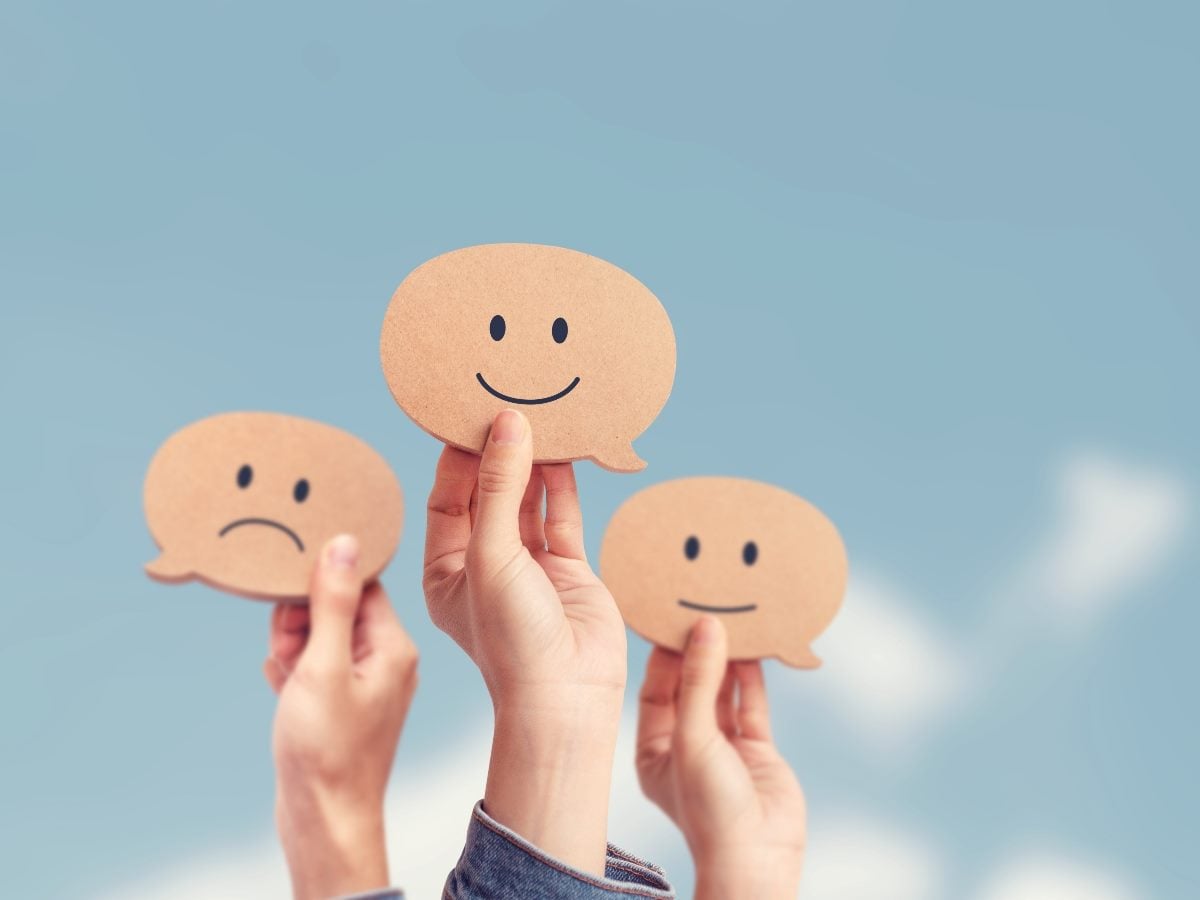 Three hands hold up speech-bubble signs displaying different customer sentiments — happy, neutral and unhappy — against a soft blue background, illustrating customer feedback, opinions and experience measurement.