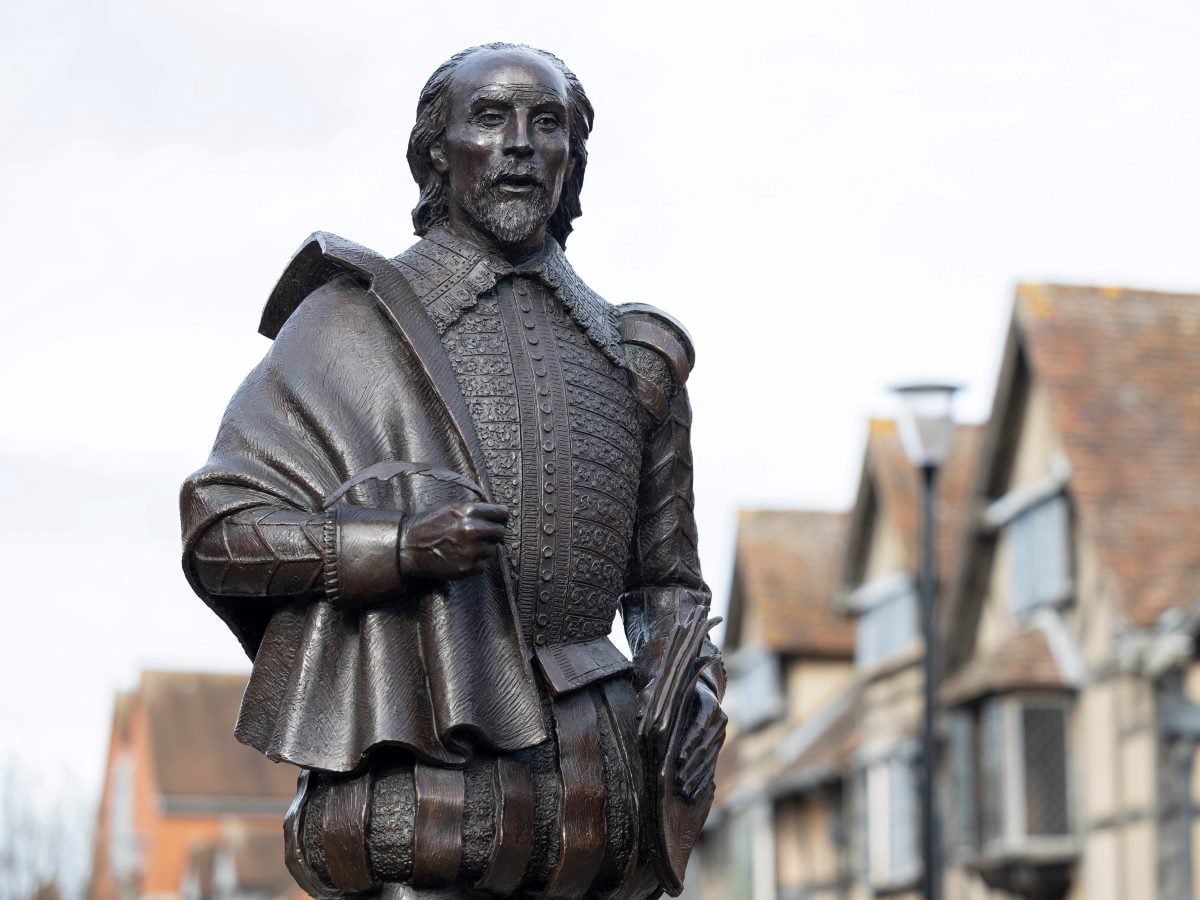 Bronze statue of William Shakespeare standing outdoors, depicting the playwright in Elizabethan attire holding papers in one hand, with historic timber-framed buildings softly blurred in the background beneath an overcast sky.