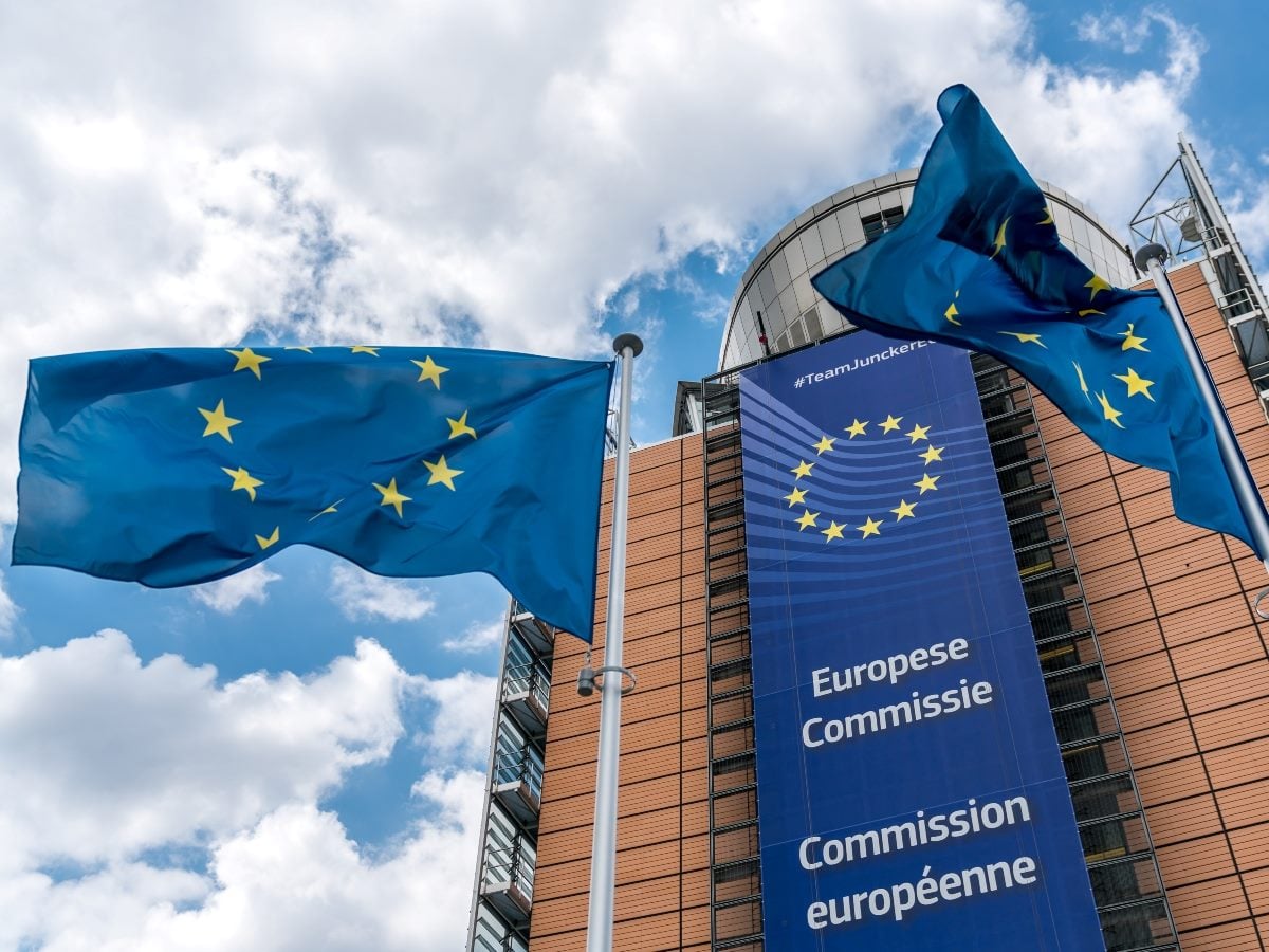 European Union flags fly outside the European Commission headquarters in Brussels, Belgium, with the commission's blue banner displayed prominently on the building beneath a partly cloudy sky.