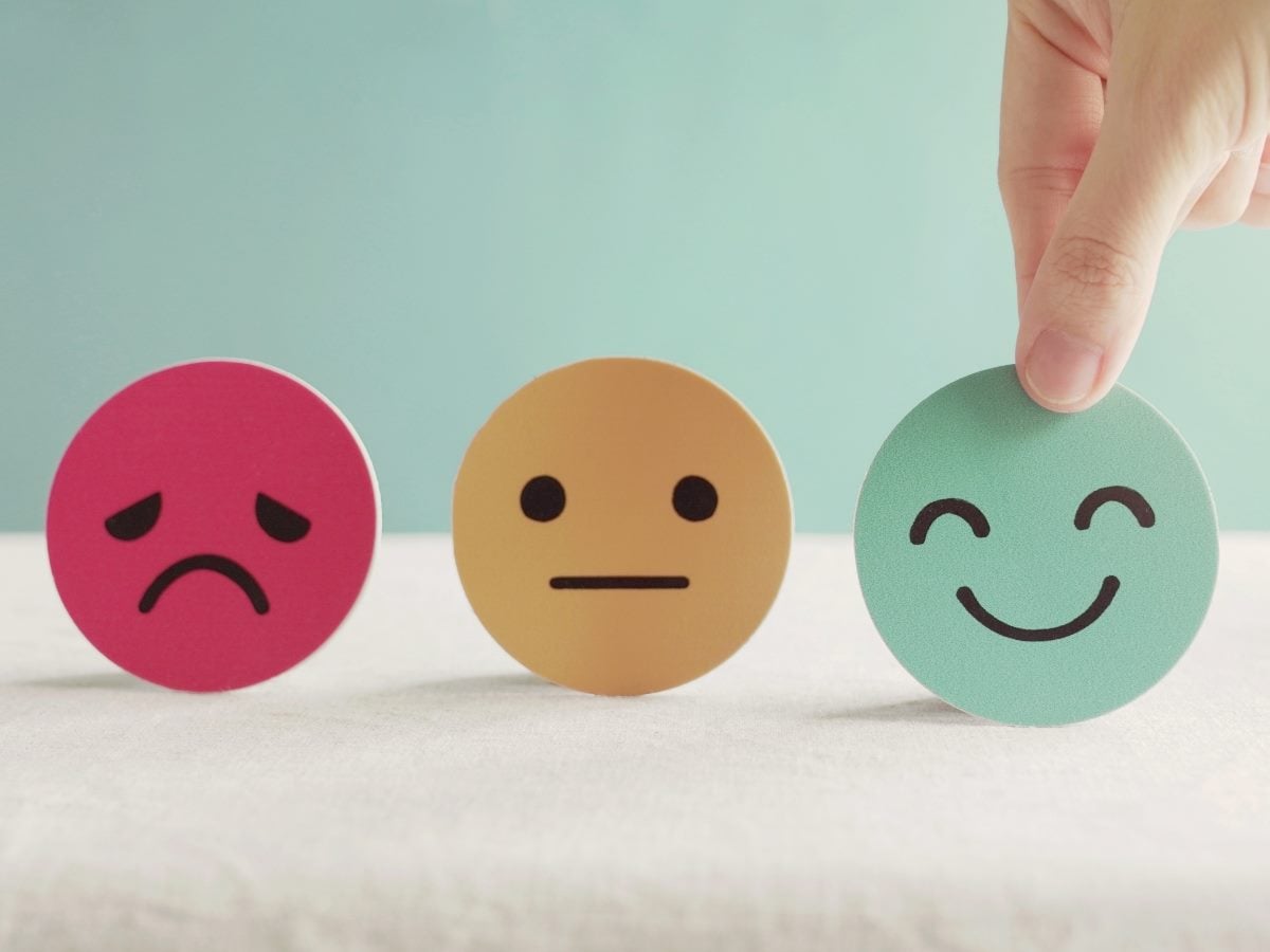 Three circular customer satisfaction icons are displayed on a white surface against a soft blue background: a red sad face, a beige neutral face and a green smiling face. A person's hand is placing the smiling icon into position, symbolizing improved customer satisfaction, positive experiences and feedback measurement.