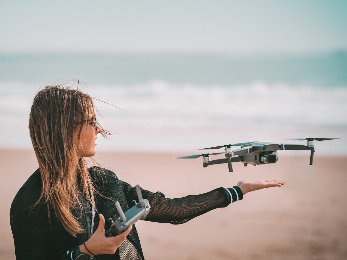 A person on a beach controlling a small quadcopter drone, holding a remote controller in one hand while the drone hovers just above their open palm with the ocean in the background.