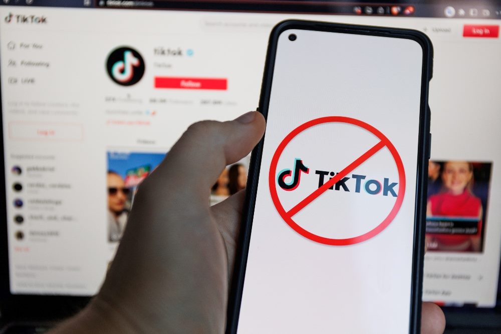 A red circle with a backslash and the TikTok logo appear on a smartphone screen while a laptop screen shows the TikTok homepage in piece about an imminent TikTok ban.