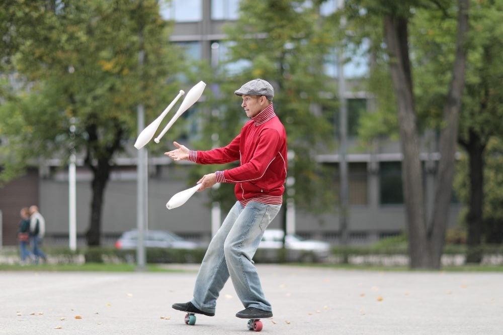 The image shows a man in a red jacket and gray cap, juggling three white clubs while standing on a skateboard. He appears focused and is performing outdoors in a park-like setting with trees and buildings in the background. There are a few pedestrians in the distance in piecer about managing an AI marketing strategy.