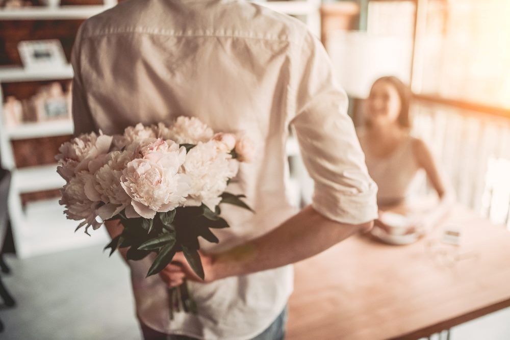 A person holds a bouquet of light pink peonies behind their back about to surprise another person sitting at a romantic cafe table in piece about marketing promotion startegy.