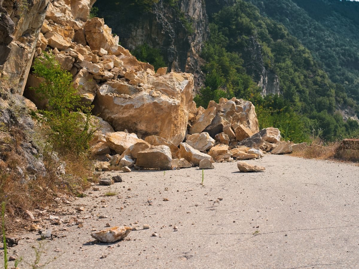 Large boulders and debris from a rockslide blocking a narrow mountain road surrounded by steep cliffs and greenery.