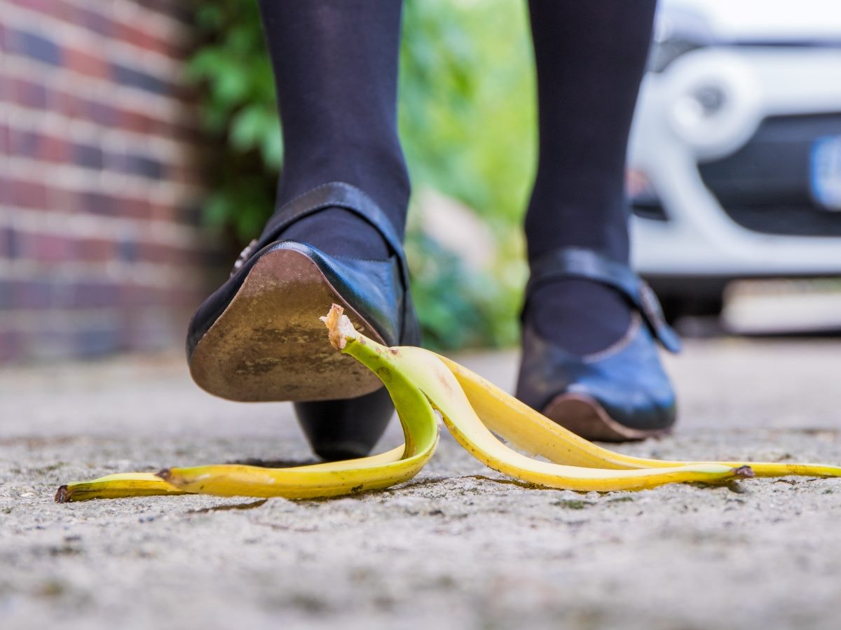 A person wearing dark shoes and tights is about to step on a banana peel on a sidewalk, captured close-up from ground level.