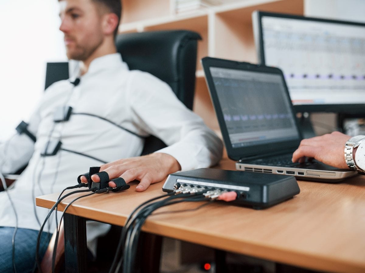 Man seated in an office setting undergoing biometric testing with sensors attached to his fingers and torso while a researcher monitors data on a laptop and external device