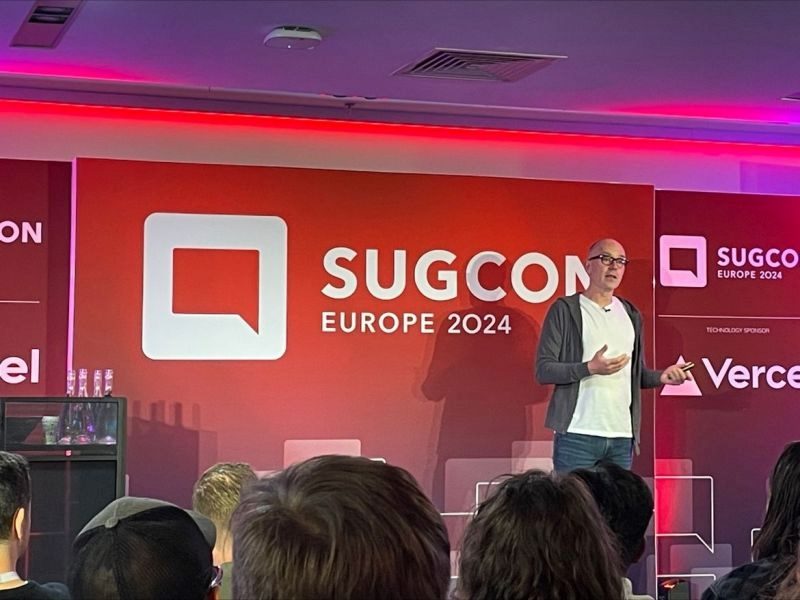 Sitecore CEO Dave O'Flanagan presenting at the SUGCON Europe Conference 2024 with the Sitecore name and a red background and the audience visible in the foreground in piece about Sitecore leadership and O'Flanagan's appointment as CEO