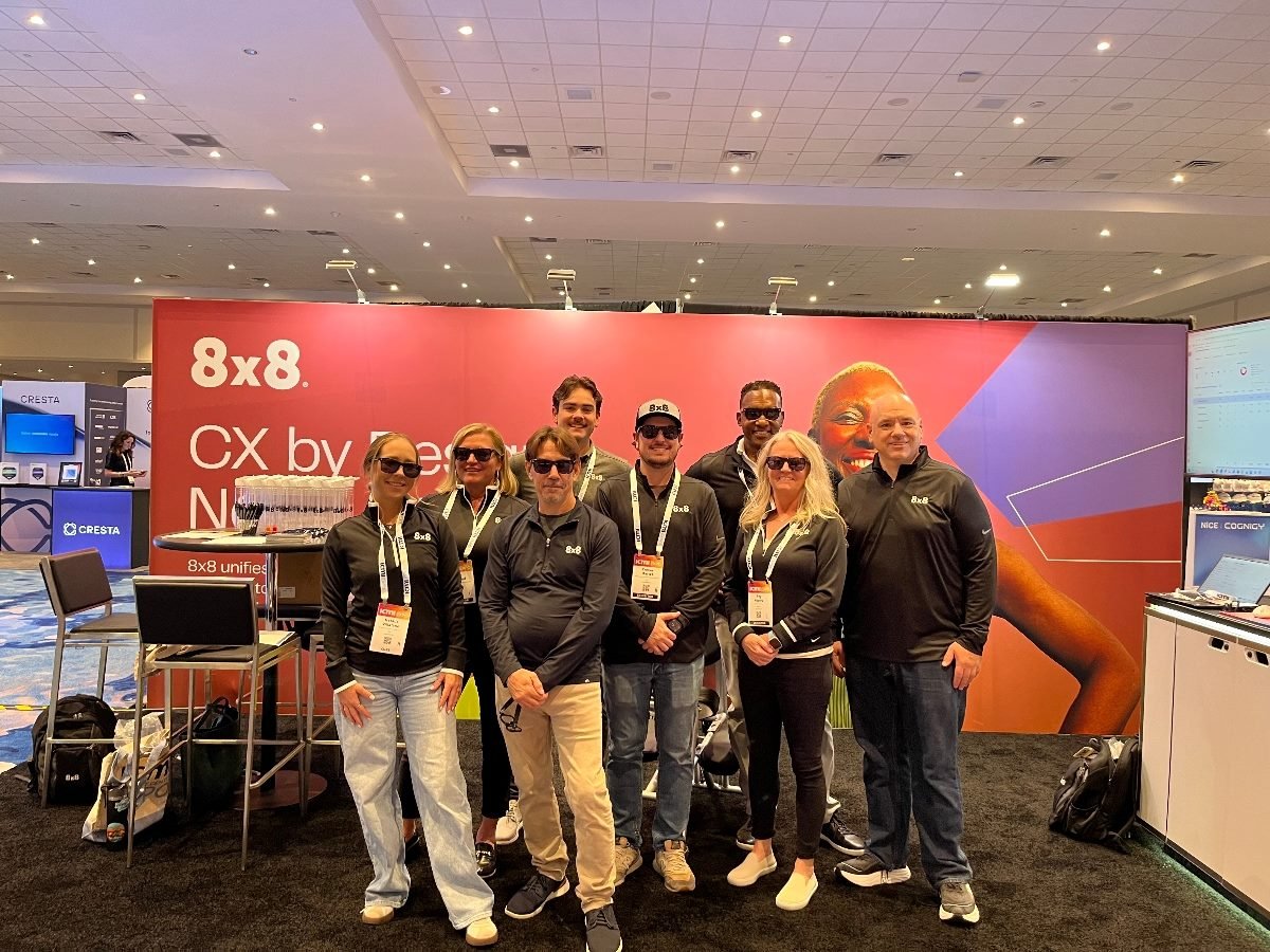 A group of eight 8x8 team members wearing black branded tops pose together at their booth during the ICMI event. The booth features a large red 8x8 backdrop with CX messaging, and the team smiles in front of it, some wearing sunglasses. High-top tables, event badges, and neighboring vendor booths are visible in the convention hall.
