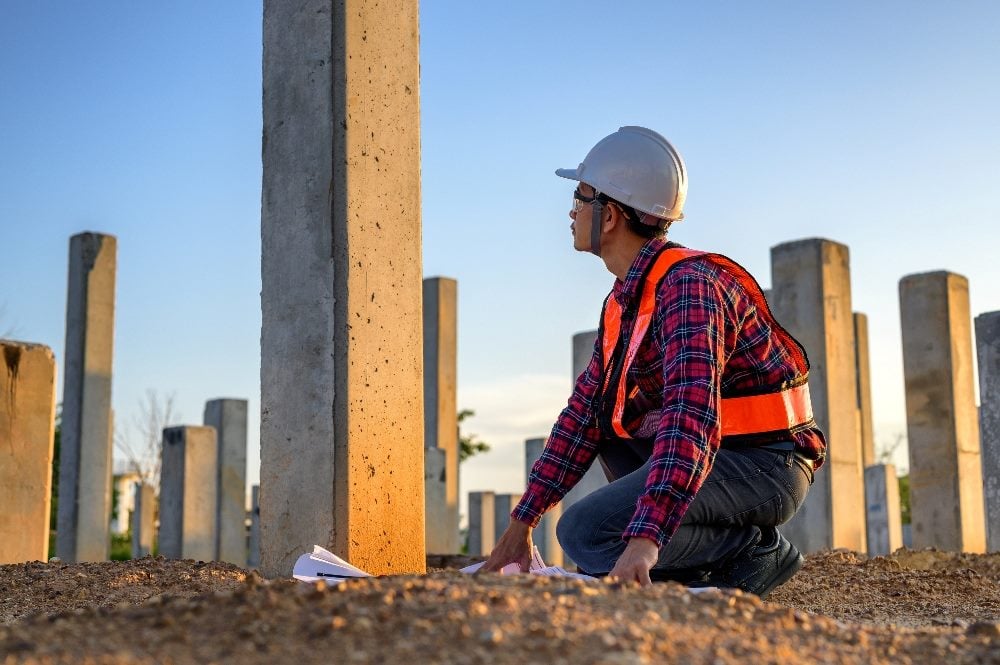 A construction engineer wearing a safety hat and flannel shirt surveys checkpoints of concrete post or piel, load-bearing posts in the ground for building a high rise in piece about customer advisory board programs and building strong foundations.