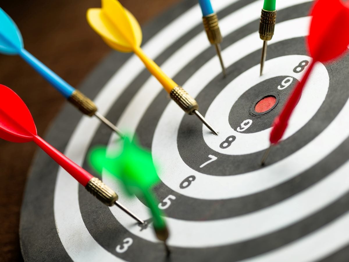 A close-up of a dartboard with several colorful darts clustered near the bullseye, including red, yellow, blue and green darts, with one red dart landing just outside the center red ring while others land closer to the middle, set against a blurred dark wooden background that keeps focus on the scattered, uneven grouping of throws.
