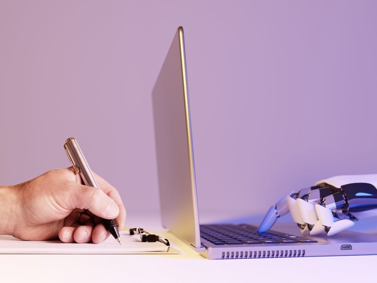 A human hand holding a pen writes on a clipboard beside an open laptop, while a robotic hand rests on the keyboard from the opposite side. The image symbolizes collaboration between humans and artificial intelligence, blending human judgment with machine-driven support in digital work environments.