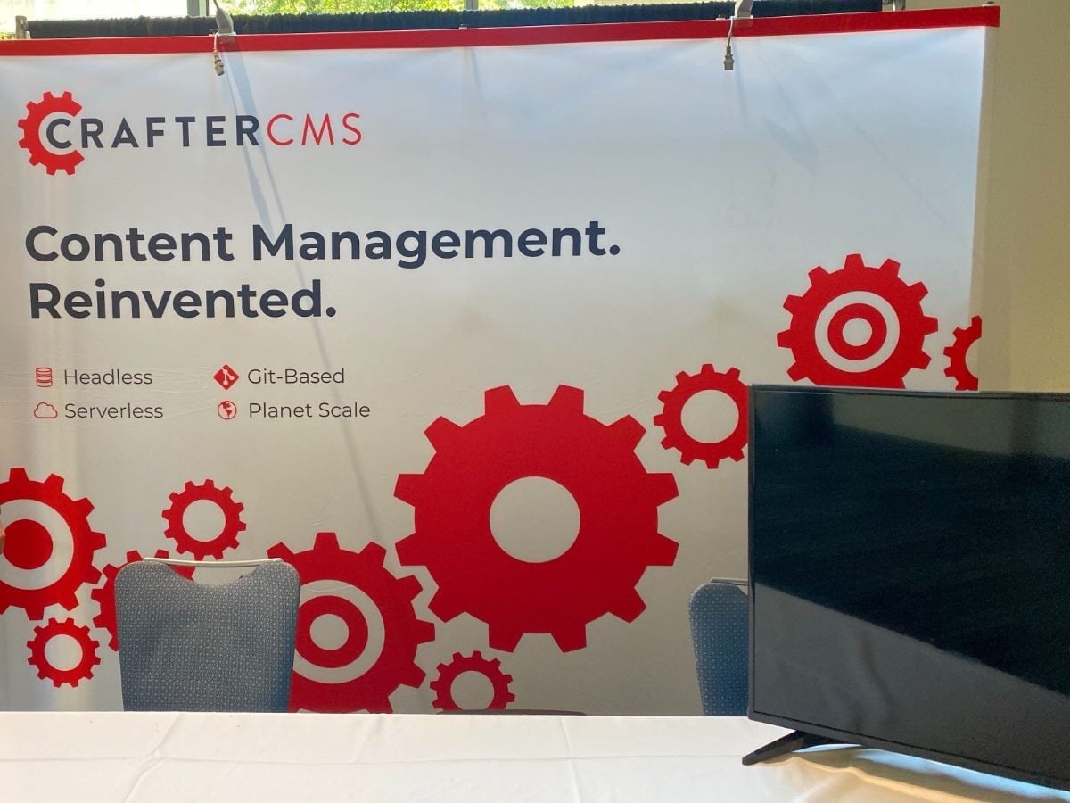 CrafterCMS booth backdrop featuring the slogan “Content Management. Reinvented.” with red gear graphics and icons highlighting headless, Git-based, serverless and planet-scale capabilities; a table, chairs and a monitor sit in front of the display.