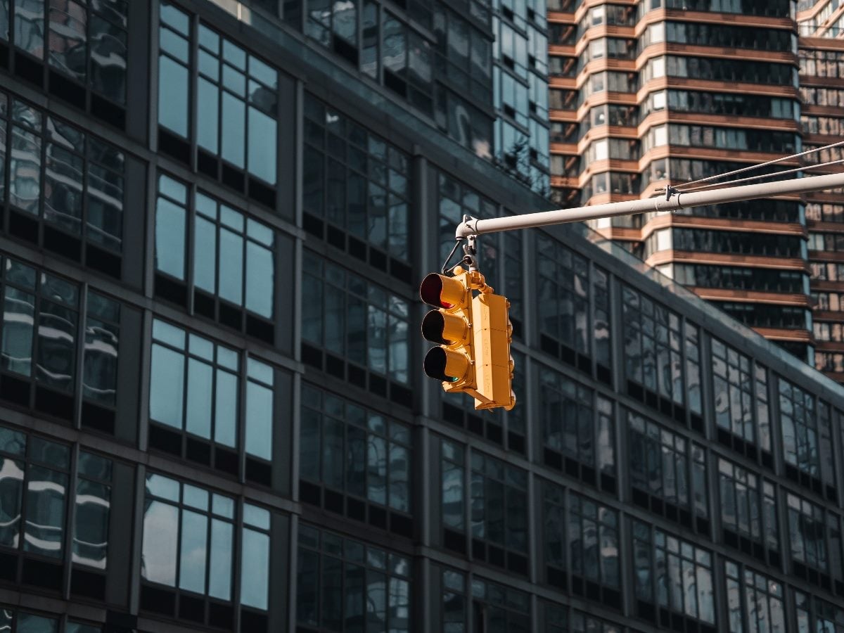 A yellow traffic signal suspended over a city street, framed against glass office buildings, symbolizing pause, decision points and controlled flow in an urban environment.