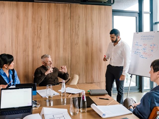 A marketing team discusses strategy in a modern office with reporting board visible to the right, representing the need for effective marketing reporting. 