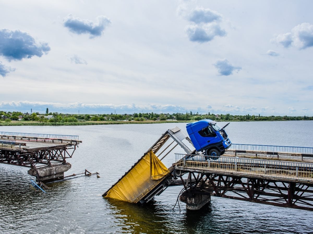 A large blue truck hangs precariously on a collapsed bridge section over a wide river, with twisted metal and a yellow tarp visible as the broken structure sinks into the water.