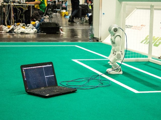 Robot with a power cord connecting to a laptop on a soccer turf in front of the net.
