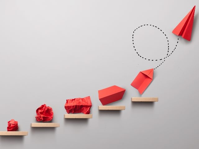 Wads of orange paper atop small wooden blocks are progressively transformed into a paper airplane with a dotted looping line from the last wad to the finished airplane, suggesting the path or change leadership in business transformation.