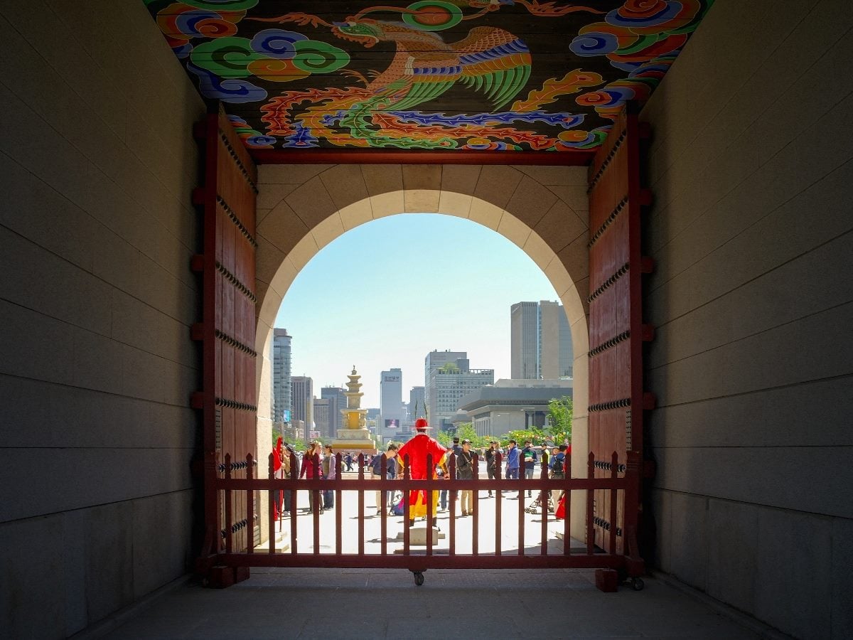 View through a large ornate palace gate with colorful painted ceiling artwork, opening onto a busy city plaza where visitors gather beneath modern skyline buildings in the background.