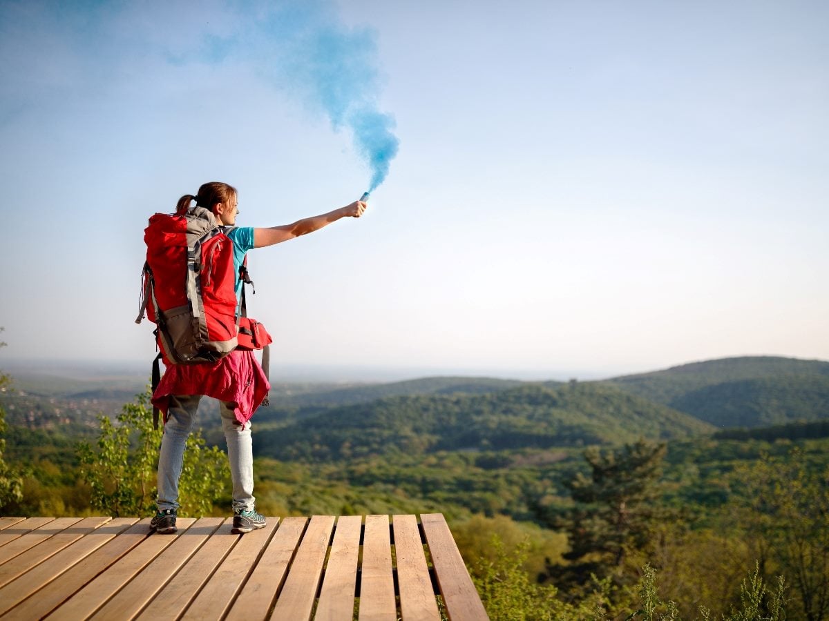 A hiker wearing a backpack stands on a wooden overlook, holding up a blue smoke flare as a signal while looking out over a wide, green mountain landscape under a clear sky.