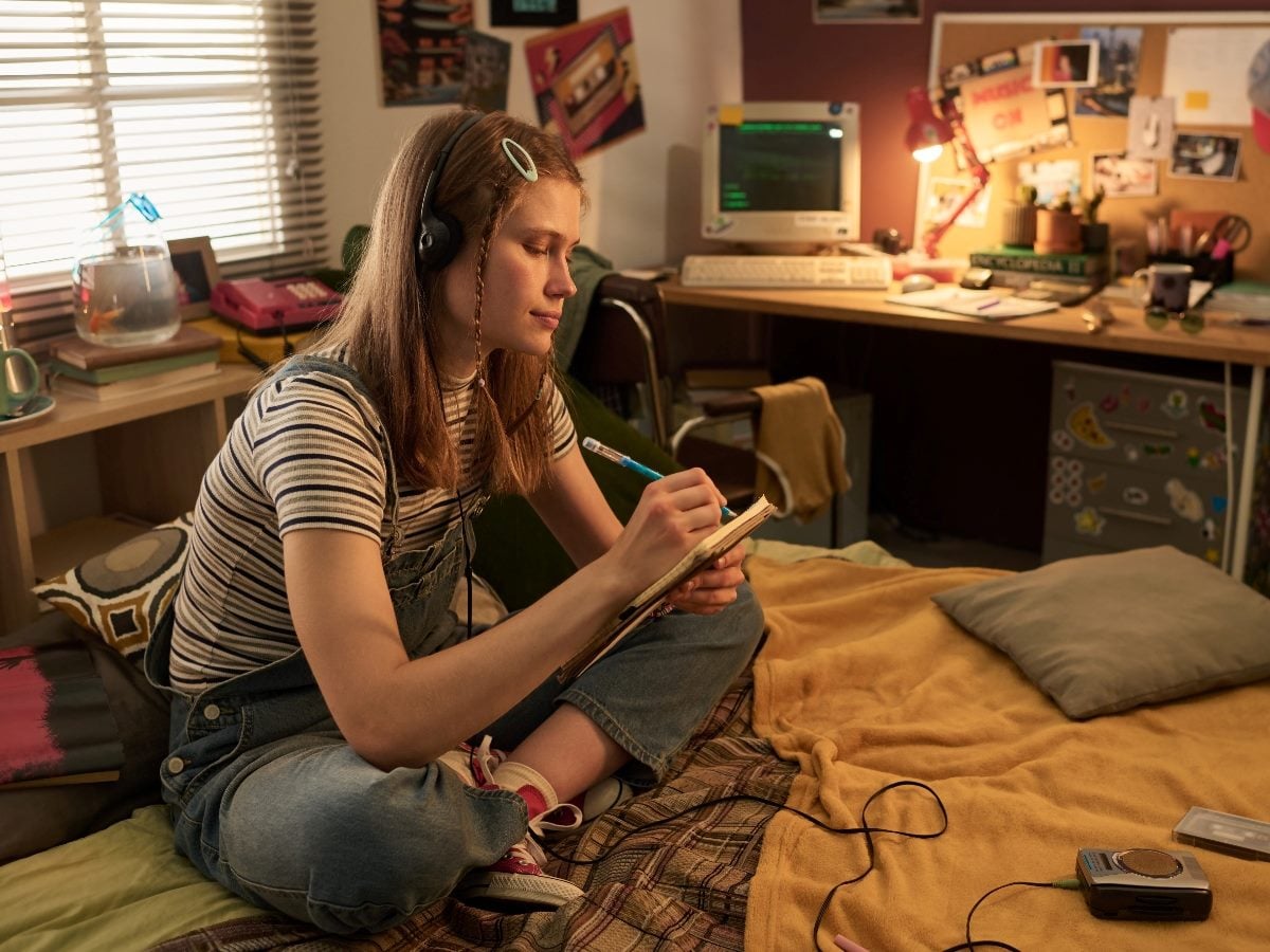 Teenager sitting cross-legged on a bed in a 1990s-style bedroom, writing in a notebook while wearing headphones, with a cassette player and vintage computer on a desk in the background.