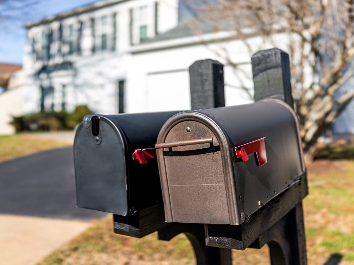 Two residential mailboxes mounted side by side on a wooden post in a suburban neighborhood. The mailbox doors are closed, with red flags lowered, and a house is softly blurred in the background.