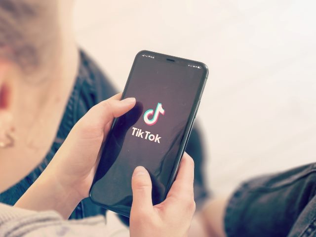A young woman sits with smart phone with TikTok logo showing, symbolizing the TikTok ecommerce launch.
