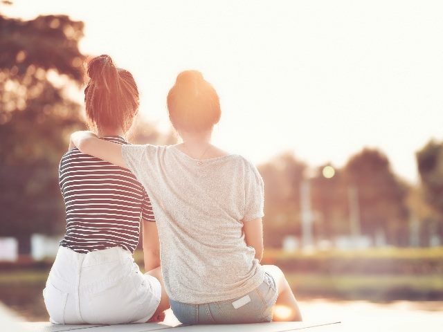 Two women resting in a garden, one with her arm around the other, watching the sunshine together. 