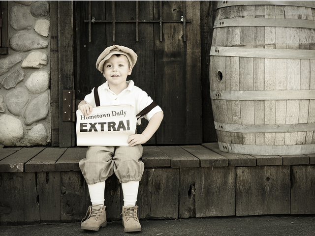 A young boy sitting down holding a newspaper that says "extra" on it.