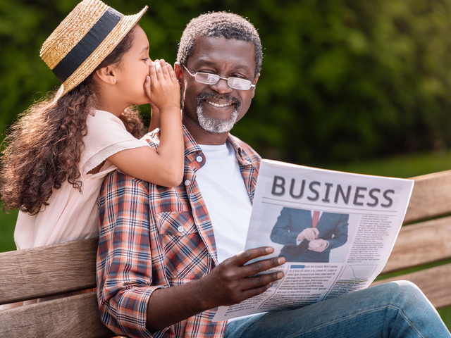 Young girl whispers into a man's ear as he sits on a bench and reads the business section of a newspaper.
