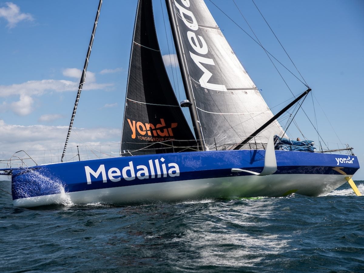 Medallia-branded racing sailboat cutting through open water, with black sails and a blue hull bearing the Medallia logo, captured mid-race under clear skies.
