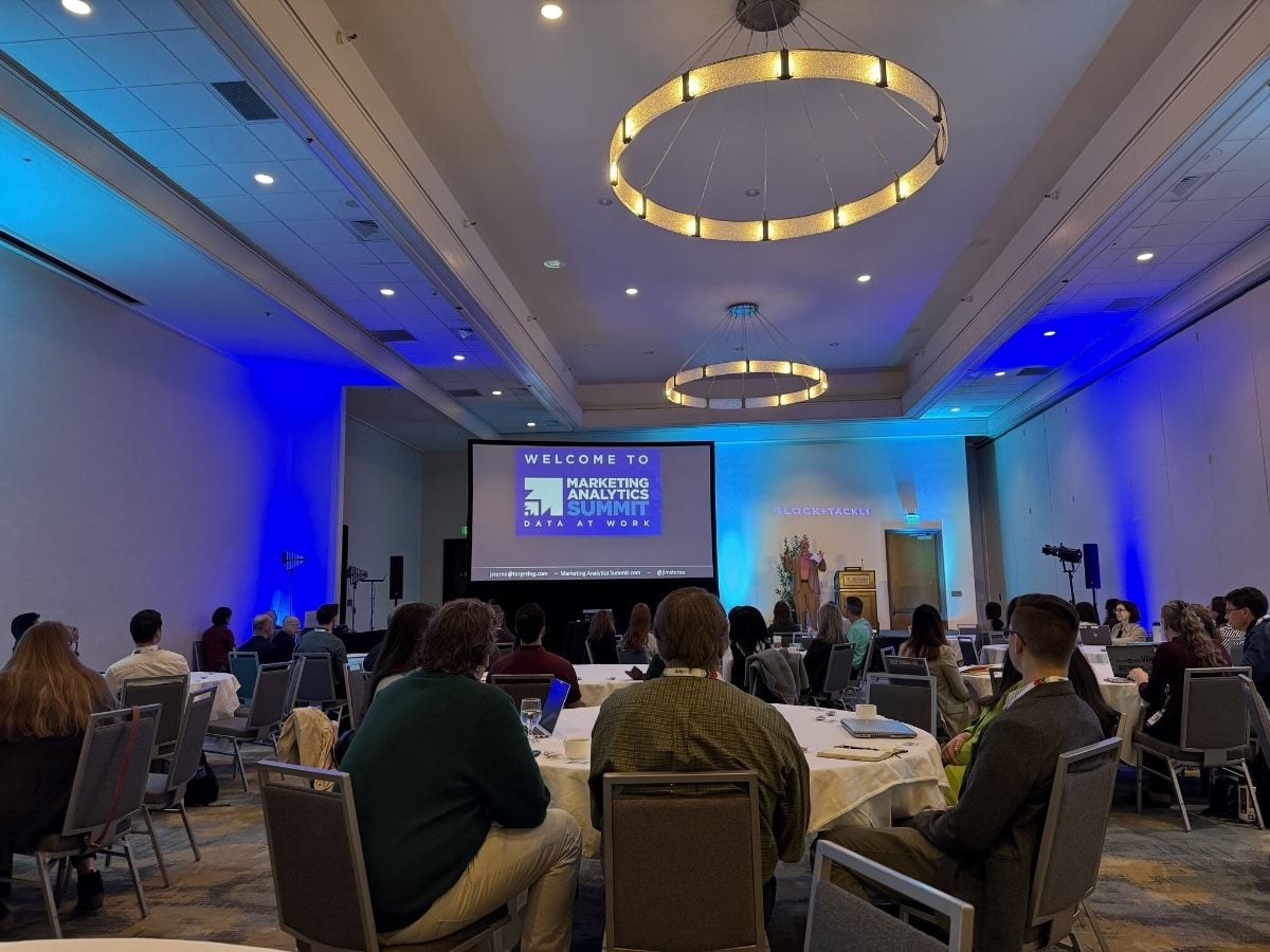 Attendees sit at round tables during a presentation at the Marketing Analytics Summit. A speaker stands near a podium at the front of a conference room while a large screen displays a welcome slide for the event. Blue accent lighting illuminates the walls, and large circular chandeliers hang from the ceiling above participants listening to the session.
