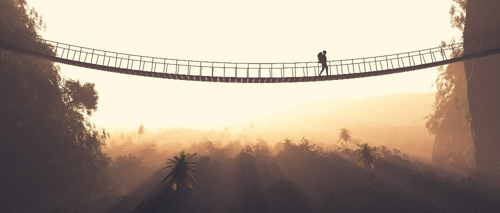 A person wearing a backpack is walking across a rope bridge above a valley with a dramatic sunset in the background in piece about social initiatives and building customer trust.